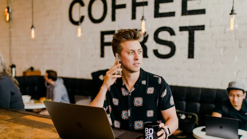 Mladý muž sedí v kavárně s notebookem, udržuje oční kontakt mimo kameru a drží šálek kávy, zatímco nápis "Coffee First" je viditelný na pozadí.