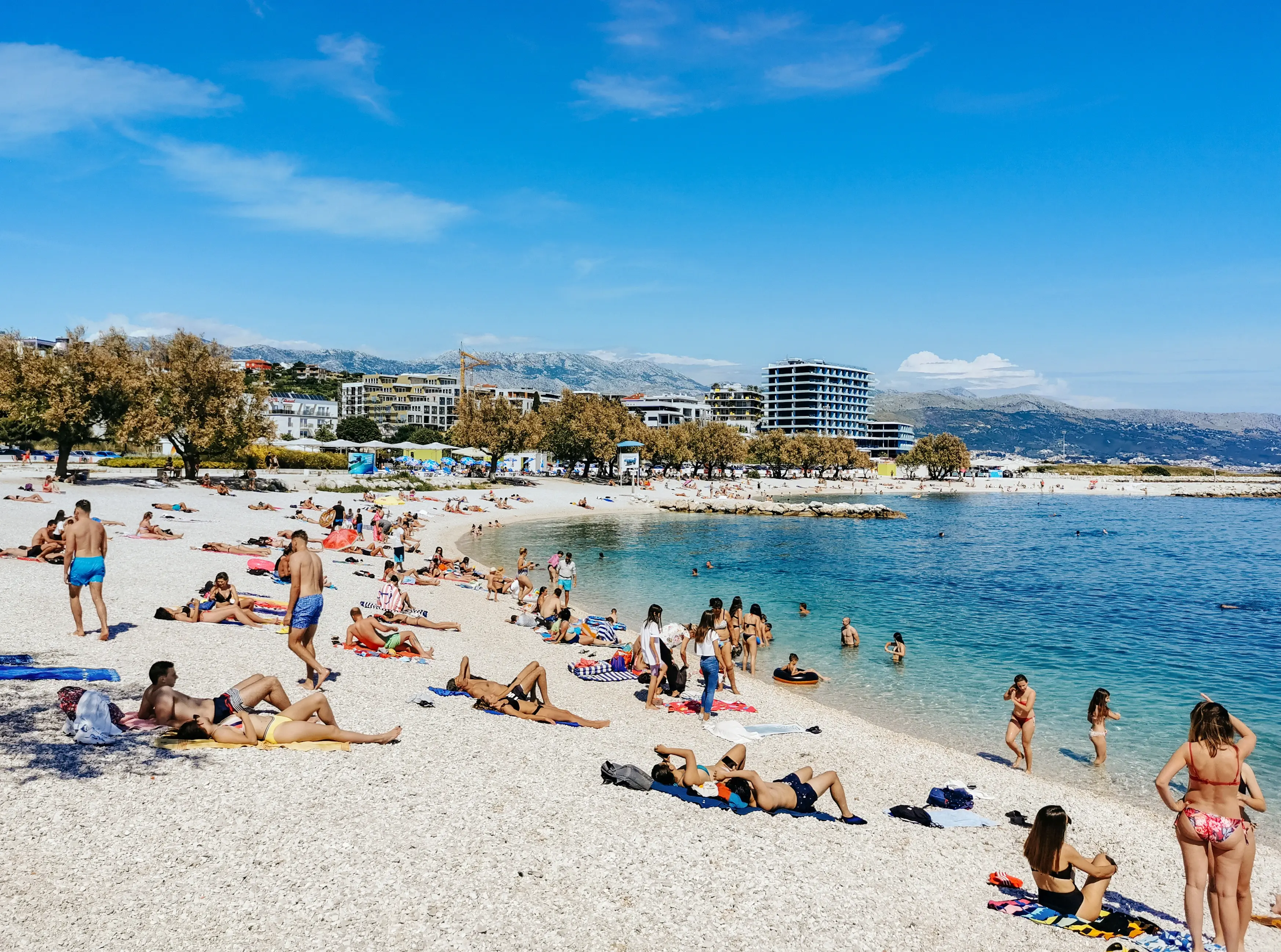 Pláž Banje v Dubrovníku plná turistů během letní sezóny, v pozadí historické hradby Starého Města.