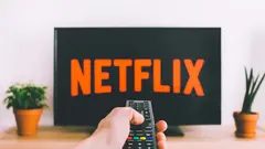 Ruka držící dálkový ovladač před televizorem s logem Netflix