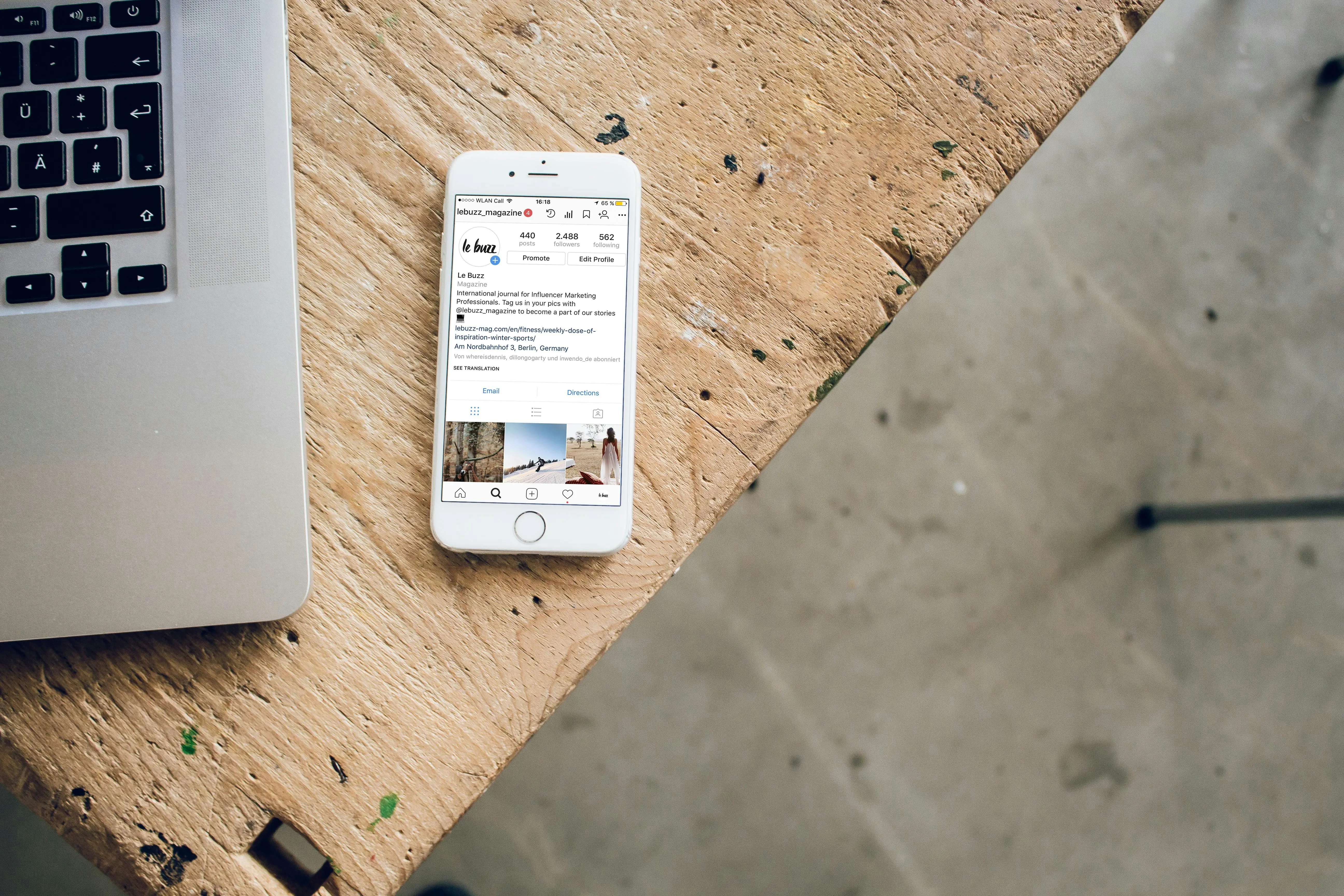 Mobilní telefon iPhone ležící na dřevěném stole vedle laptopu, zobrazující Instagramový feed.