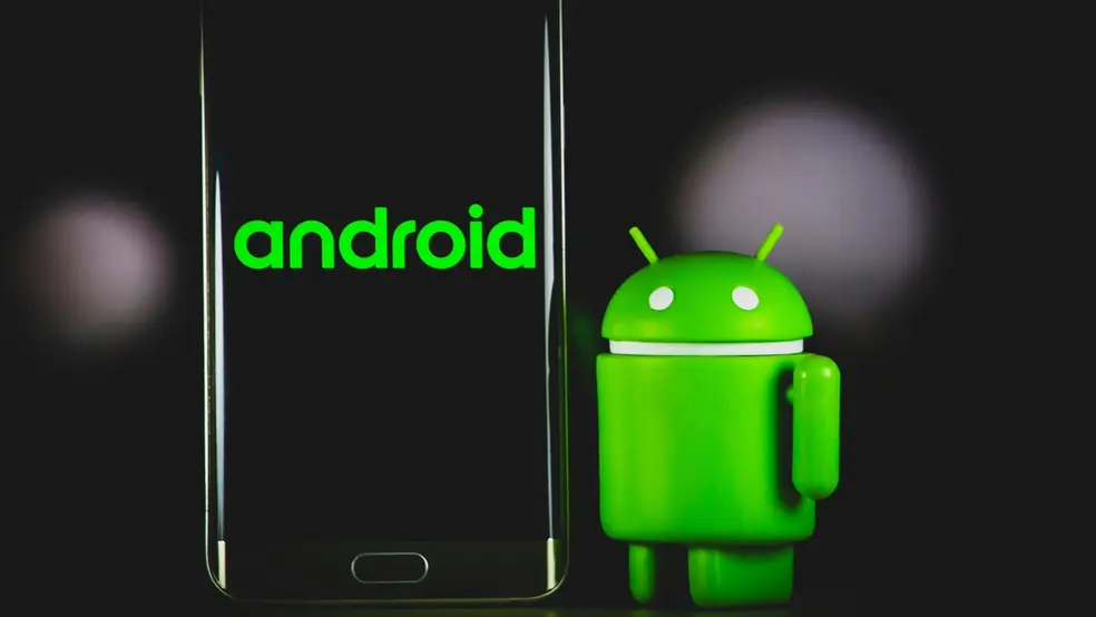 Android maskot stojící vedle smartphonu se zeleným logem Androidu na obrazovce.