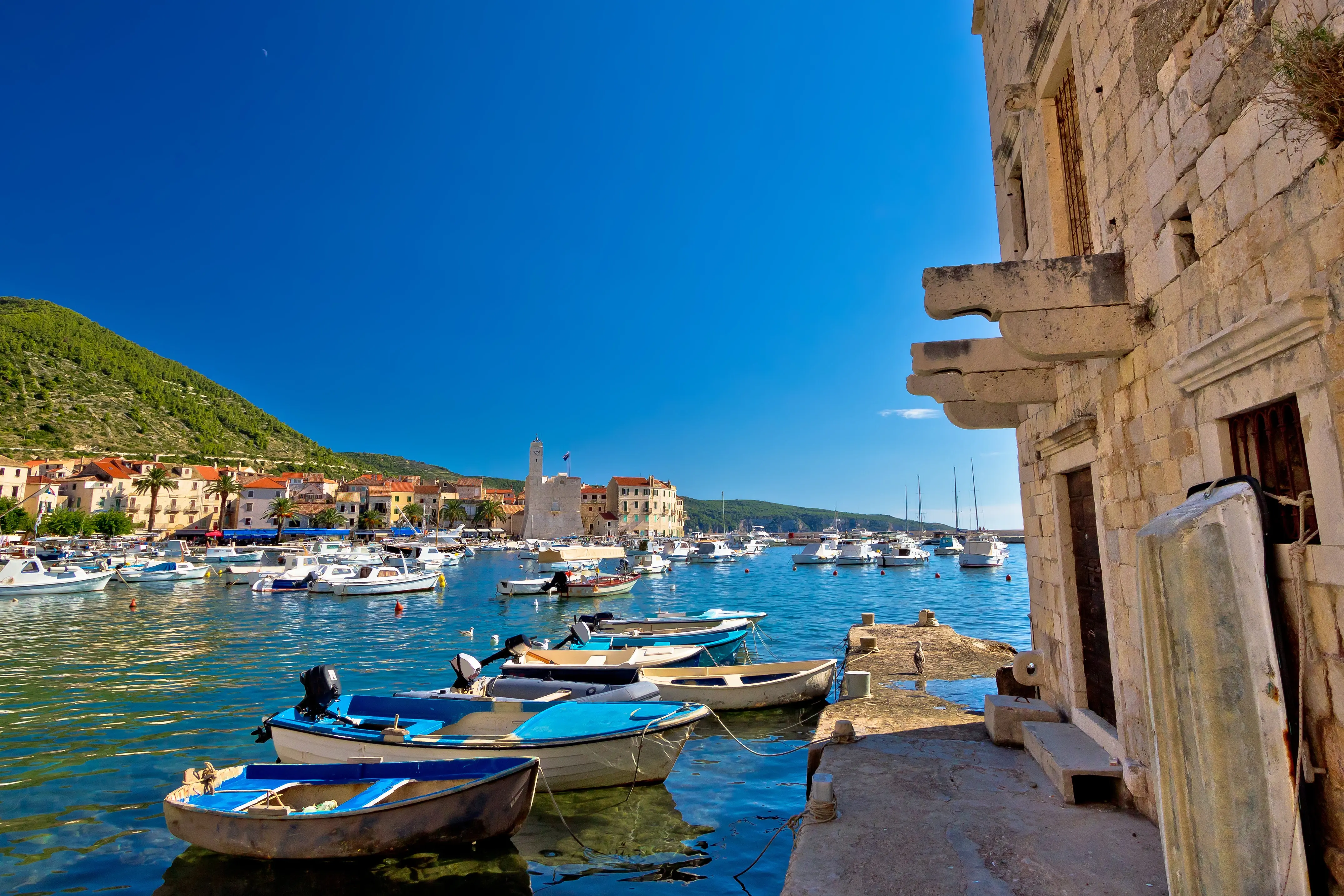 Lidé na nábřeží města Hvar lemovaném palmami s pevností v pozadí