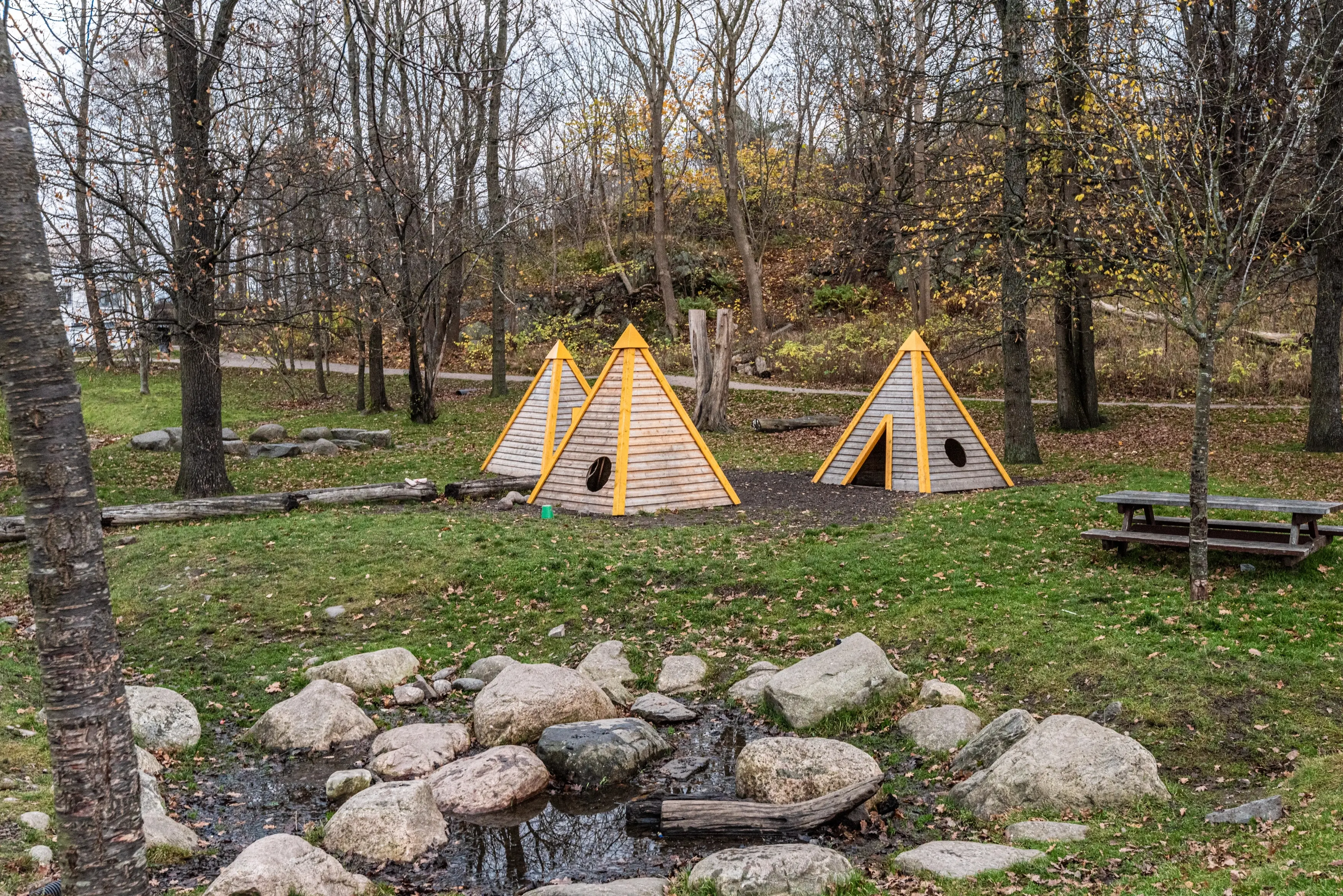 Tři dřevěné pyramidové domečky obklopené stromy, s piknikovým stolem a potůčkem v popředí