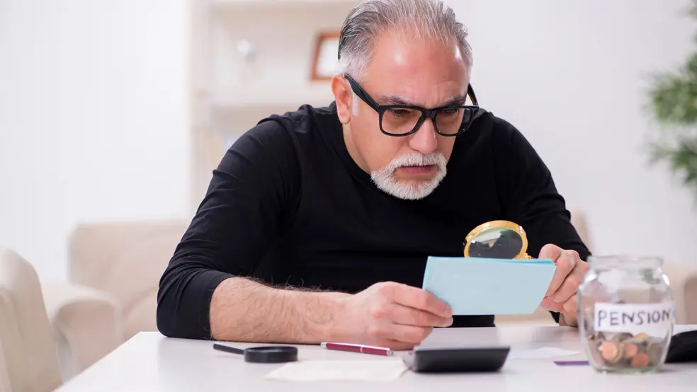 Starší muž v černém oblečení se silnými brýlemi zkoumá složenku s lupou, vedle sklenice na důchod