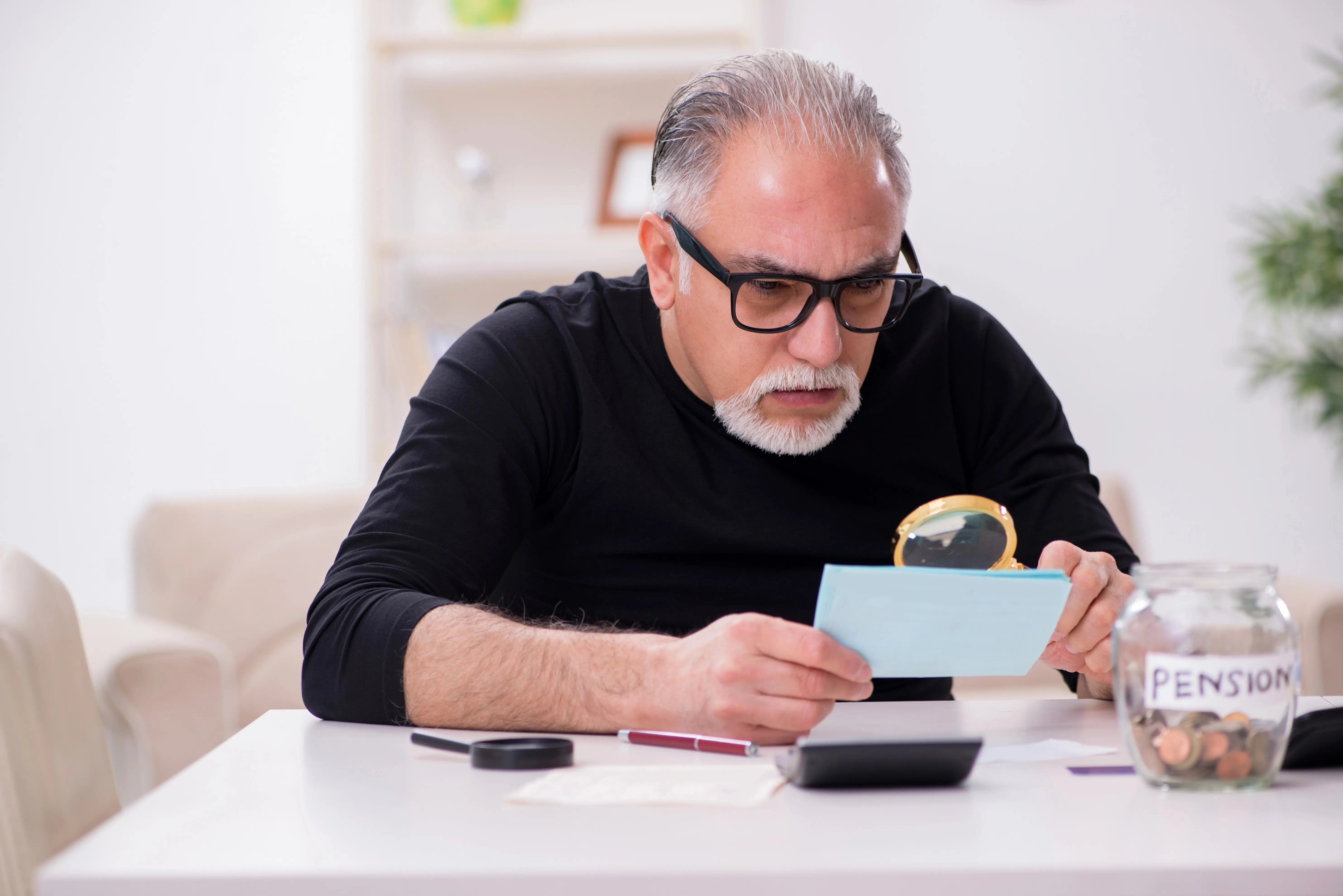 Starší muž v černém oblečení se silnými brýlemi zkoumá složenku s lupou, vedle sklenice na důchod