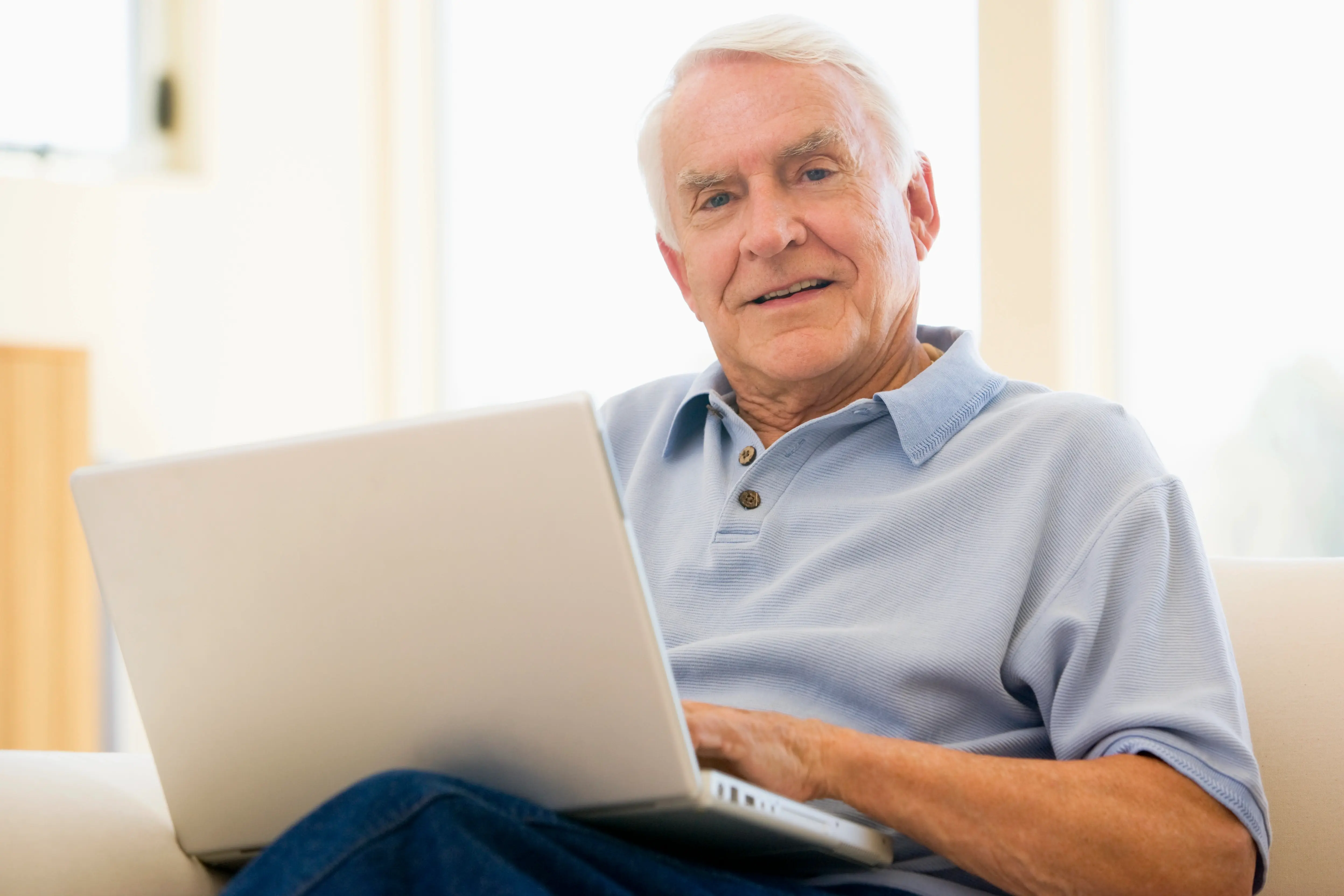 Starší muž v modrém tričku sedí na gauči a pracuje na notebooku, usmívá se do kamery