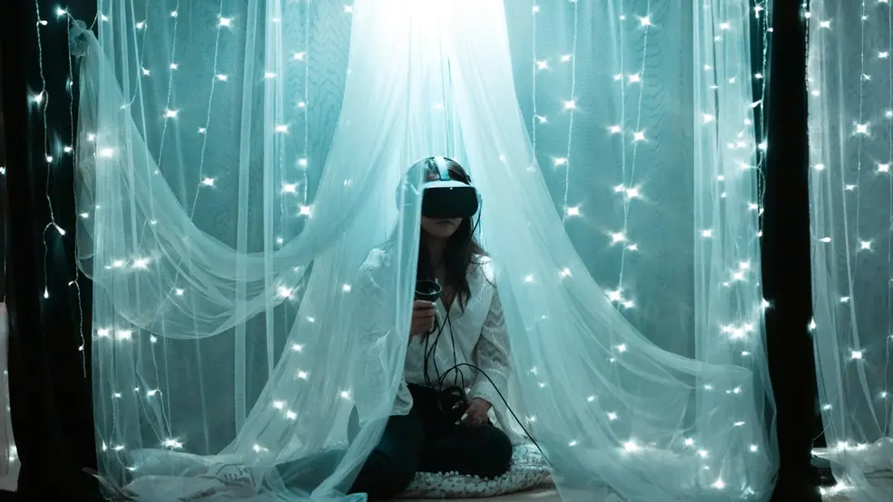 Žena s VR headsetem sedící pod bílými závěsy s rozsvícenými světýlky.