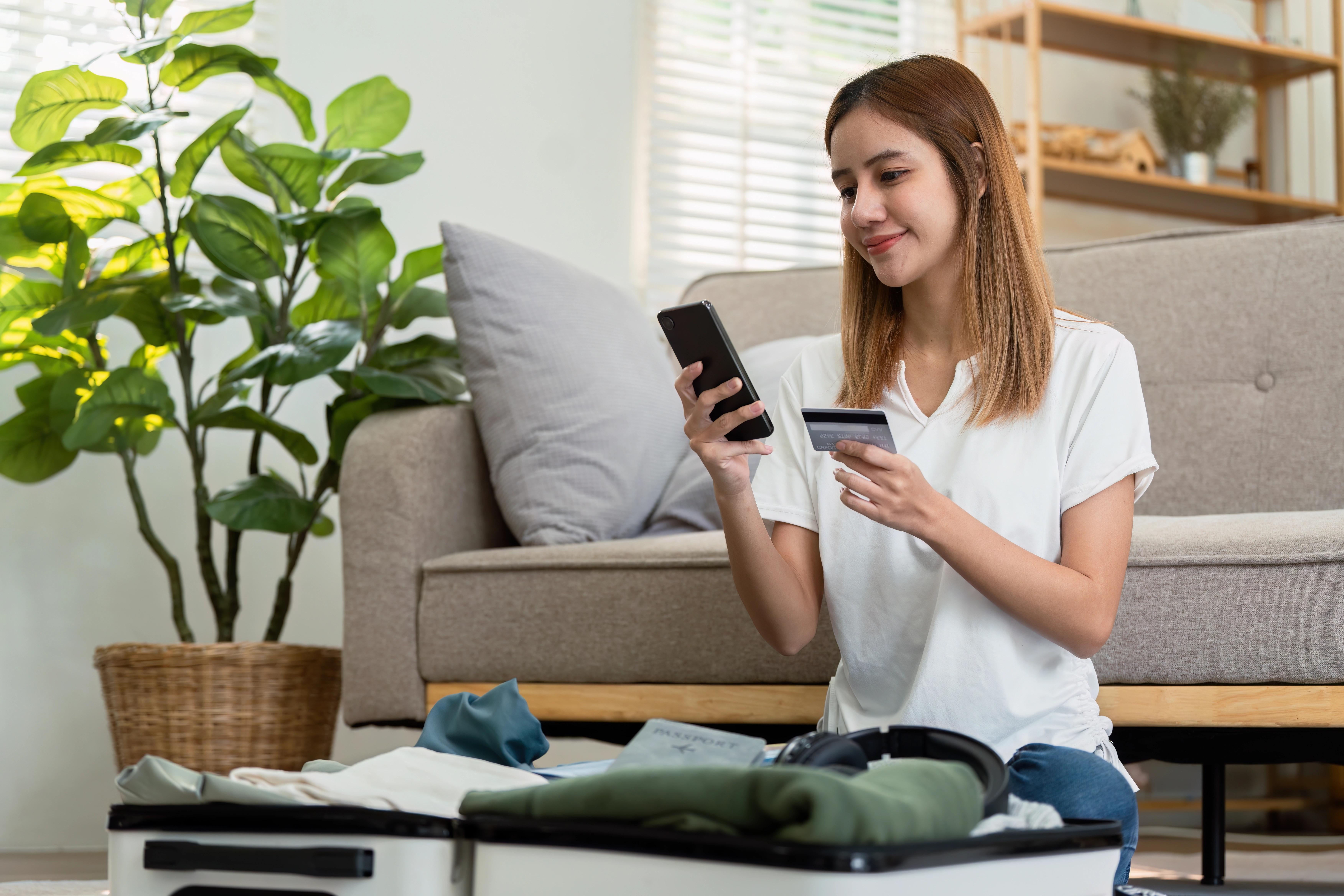 Mladá žena sedí na podlaze s telefonem a platební kartou, u sebe má kufr s oblečením