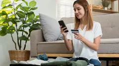 Mladá žena sedí na podlaze s telefonem a platební kartou, u sebe má kufr s oblečením