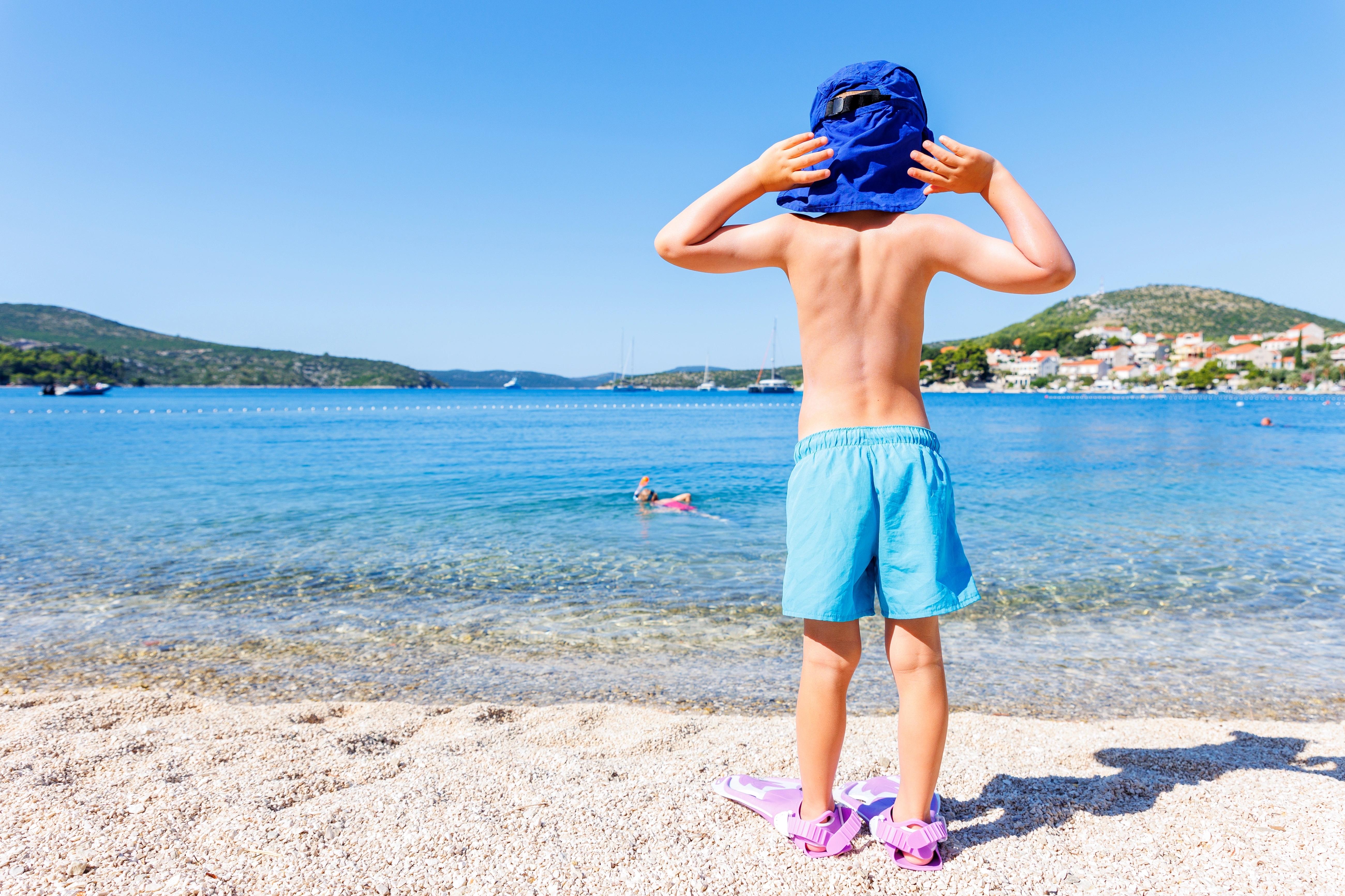 Malý chlapec v klobouku stojí na písčité pláži a dívá se na moře.