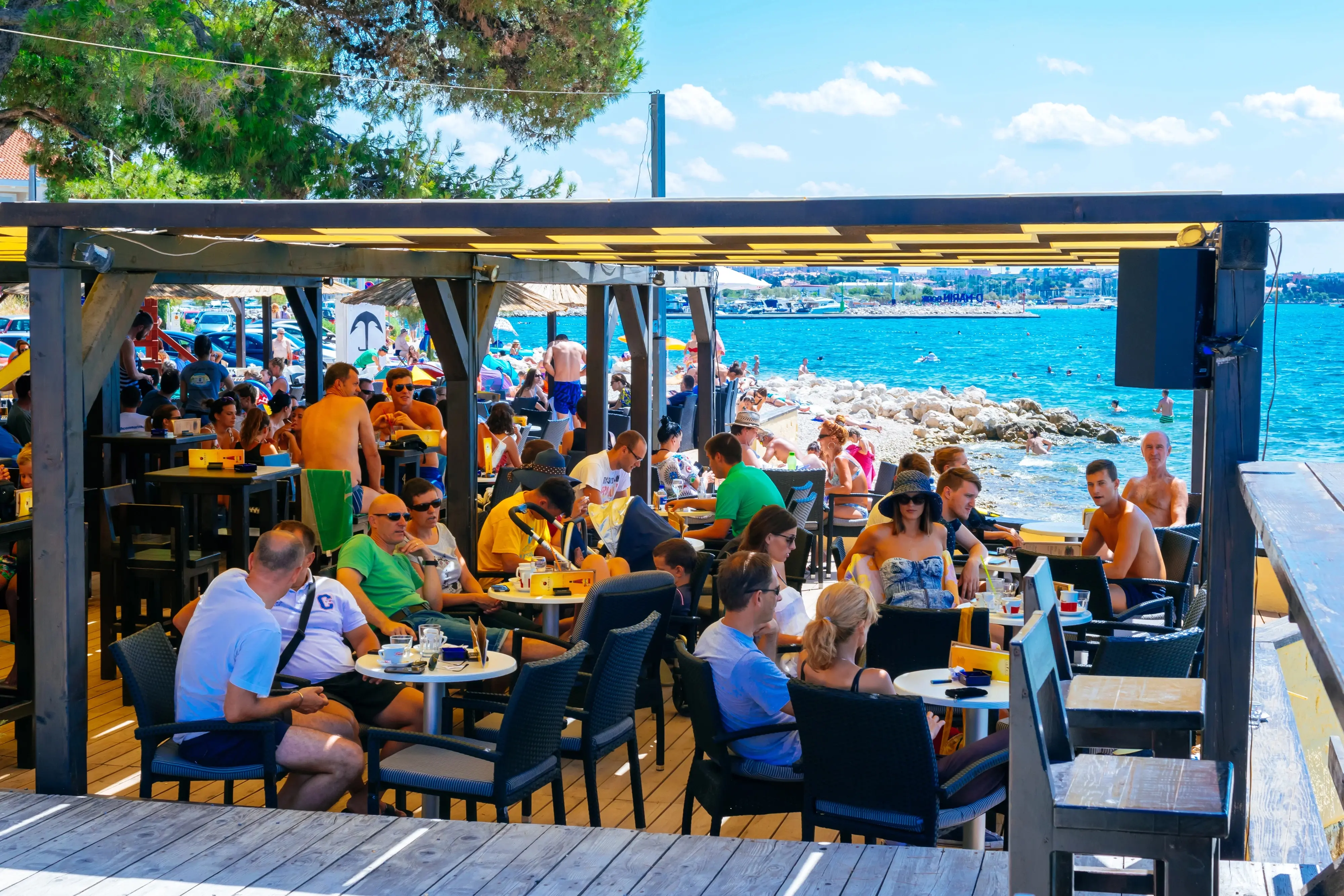 Rušný plážový bar plný lidí u moře za slunečného dne, výhled na azurové moře a kamenitou pláž