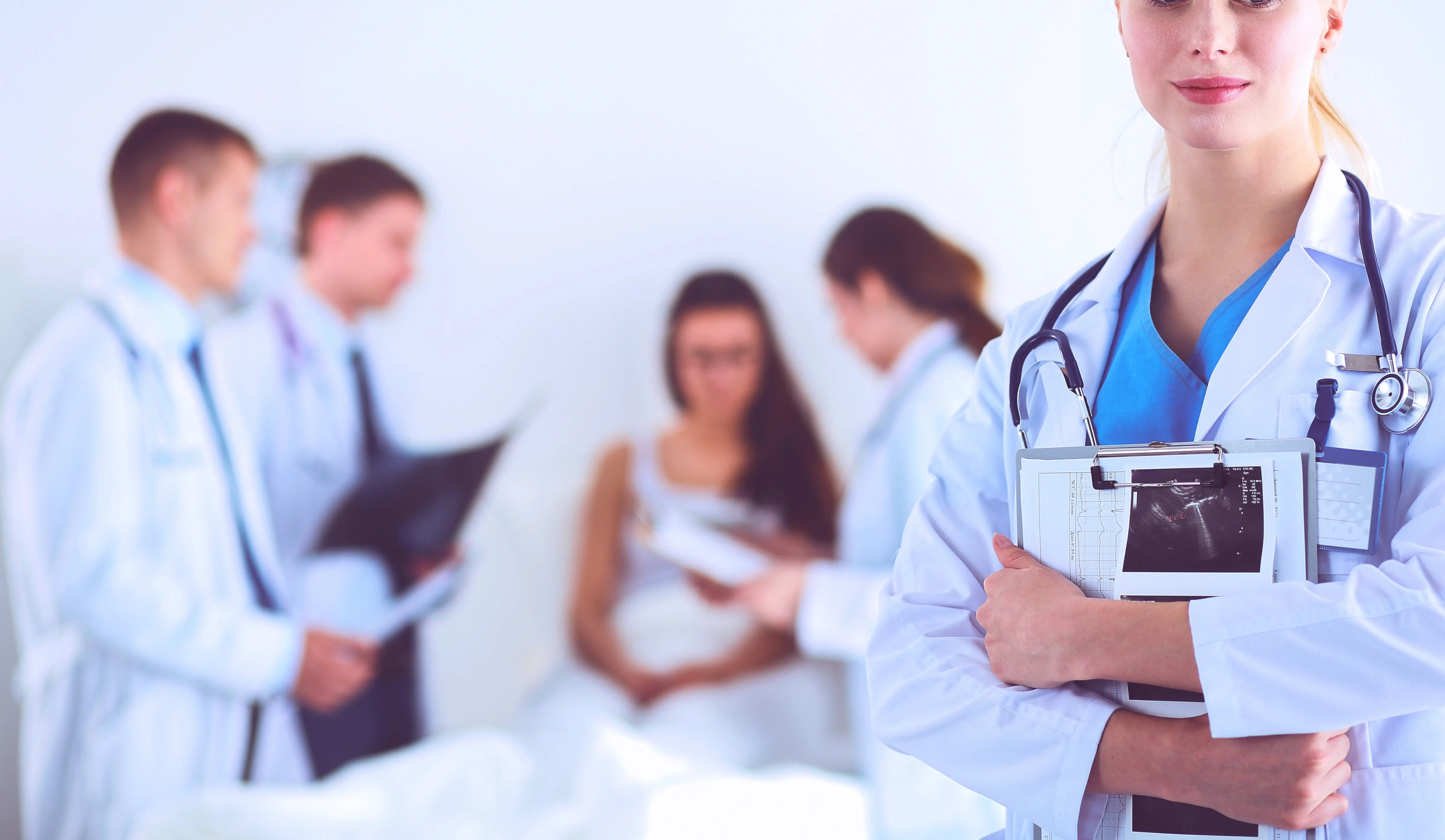 Mladá lékařka v popředí a skupina doktorů u pacientky v pozadí