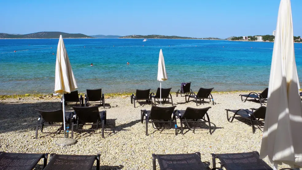 Letní pláž se slunečníky a lehátky, klidné moře v pozadí, modrá obloha a ostrovy na horizontu