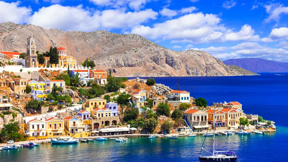 Pohled na přístav s barevnými domy a loděmi na řeckém ostrově Symi, v pozadí skalnaté hory