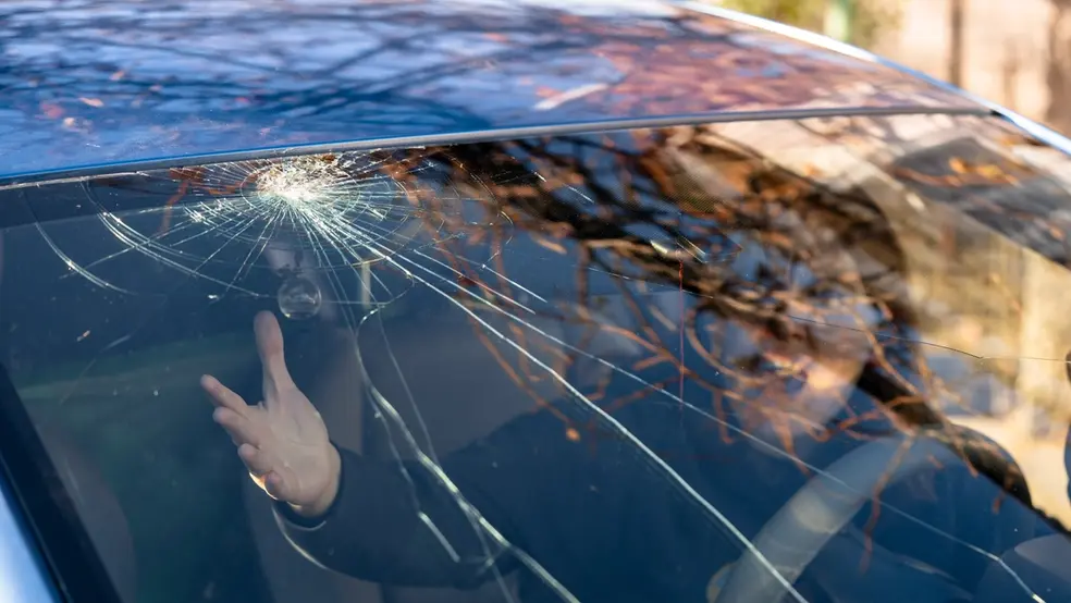 Pohled na prasklé čelní sklo automobilu, z pohledu zevnitř, s osobou uvnitř ukazující rukou na prasklinu.