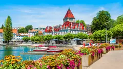 Historický zámek s červenou střechou u jezera obklopeného zelení.