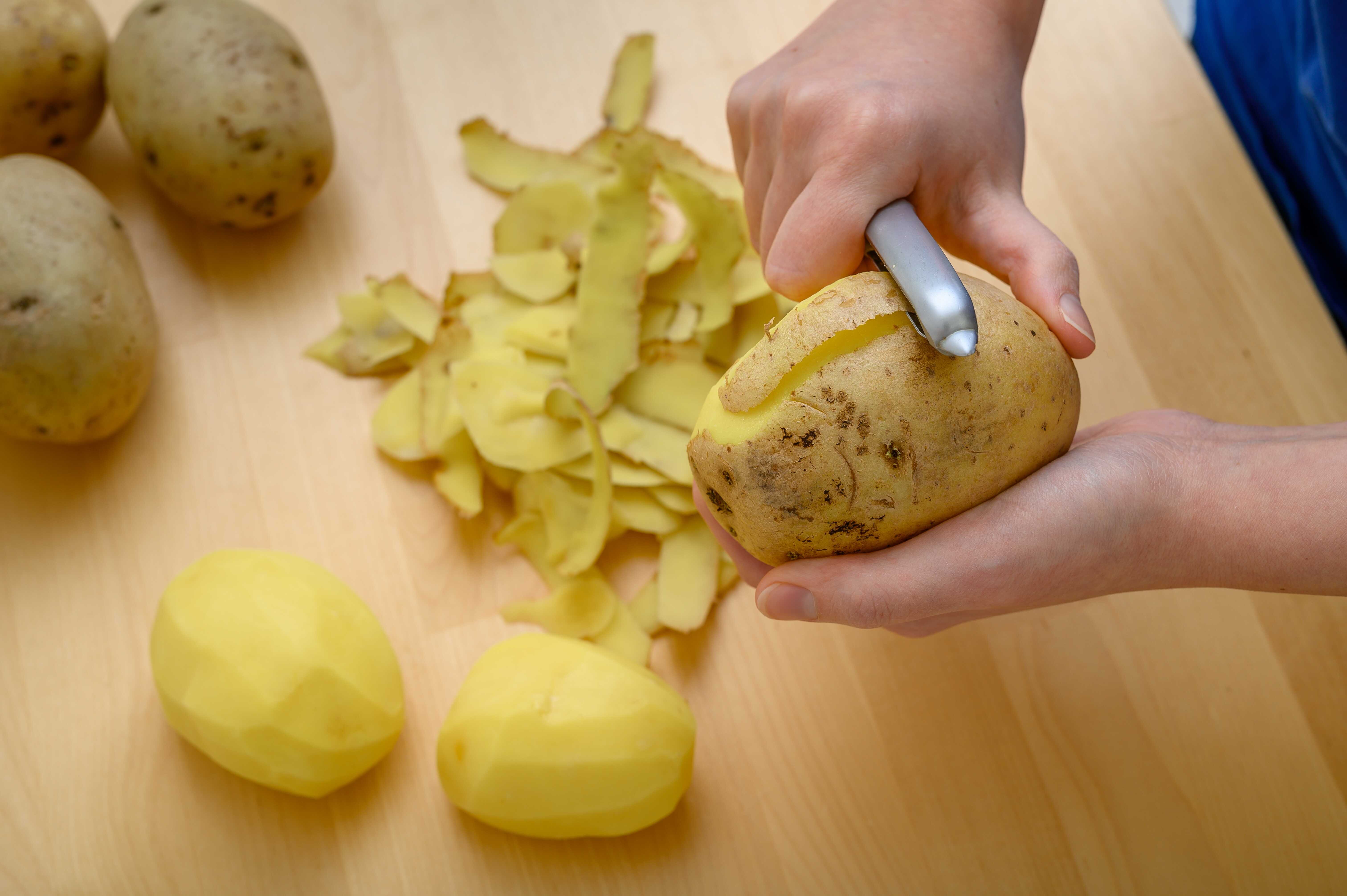 Ruka držící škrabku, kterou loupe bramboru, vedle hromádky slupek a již oloupaných brambor