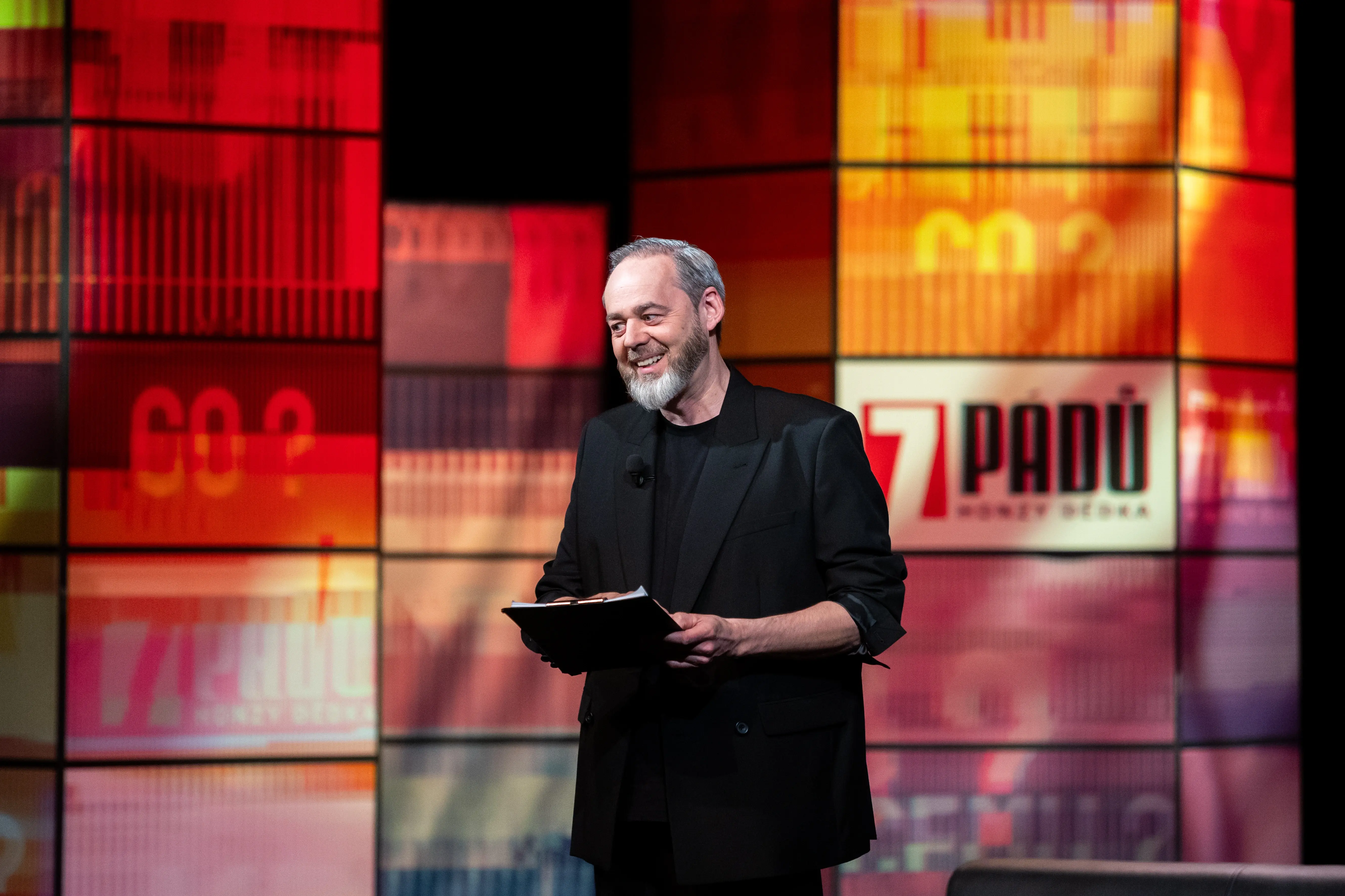 Moderátor Honza Dědek stojí před barevným pozadím talkshow a drží desky s poznámkami