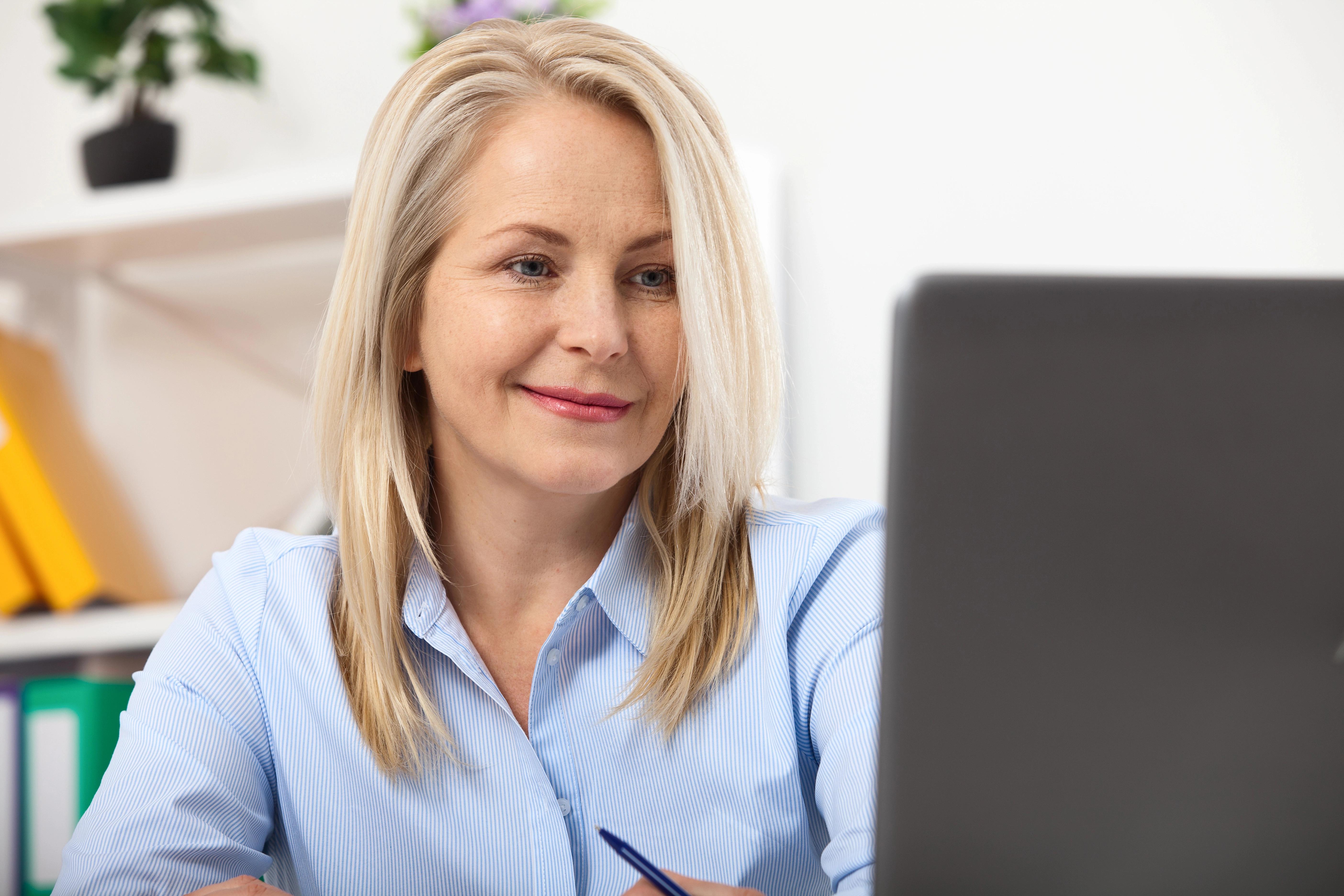 Blondýnka sedí u stolu s notebookem a usmívá se do kamery